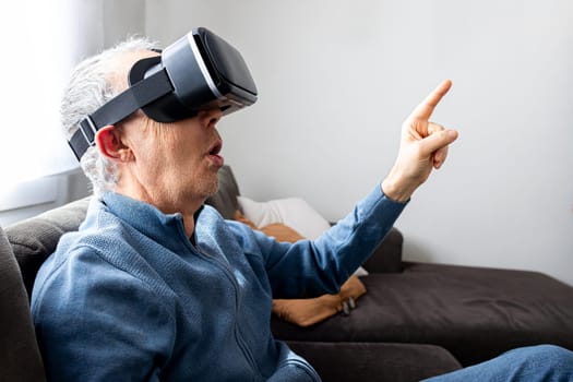 Senior man sitting on the sofa with surprise expression using virtual reality simulator at home to buy groceries. Home and technology concept.