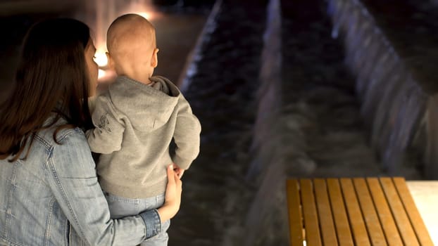 Rear view of young mother with her little son standing near the fountain in the late evening. Mom and her boy looking at the fountain.