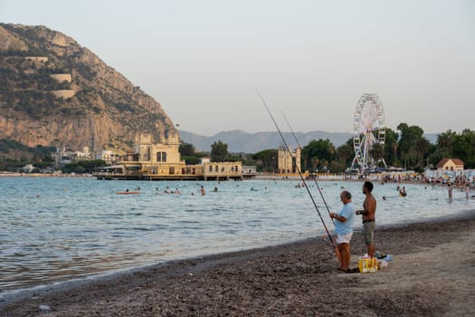 Mondello, Italy - July 17, 2023: Two men fishing with fishing rods at Mondello Beach.