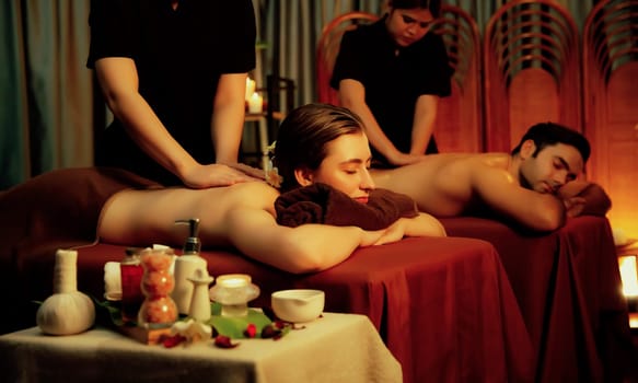 Caucasian couple customer enjoying relaxing anti-stress spa massage and pampering with beauty skin recreation leisure in warm candle lighting ambient salon spa at luxury resort or hotel. Quiescent