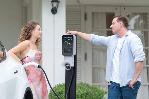 Happy and lovely couple with eco-friendly conscious recharging electric vehicle from EV home charging station. EV car technology utilized for residential home to future sustainability. Synchronos