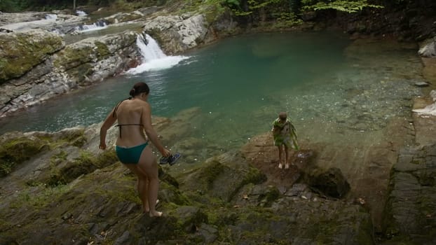 Woman with boy on pond in forest. Creative. Woman with child resting on pond with waterfall. Family is relaxing in pond with waterfall in wild green forest.