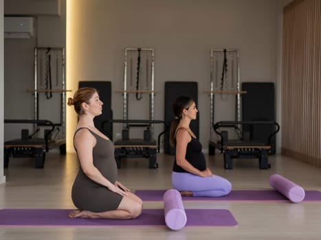 Two pregnant women are sitting on a yoga mat. Prenatal yoga