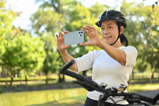 Happy middle age woman in bike helmet stops to taking photo of landscape with mobile phone
