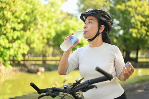 Active senior woman drinking water from a bottle after workout against blurred green park background