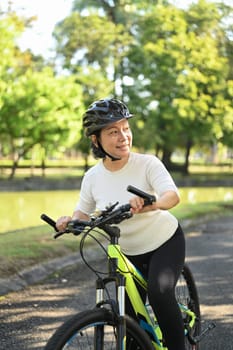 Portrait of happy retired woman in helmet riding bicycle in park on sunny summer day