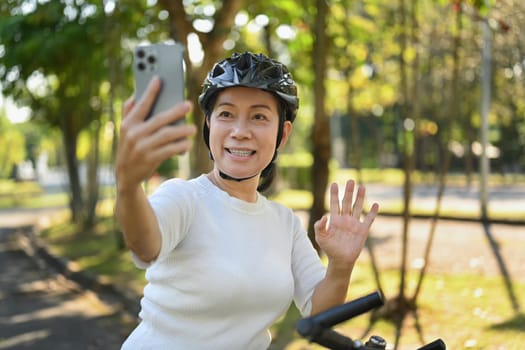 Cheerful middle age woman having video call on mobile phone while resting from cycling in a green park