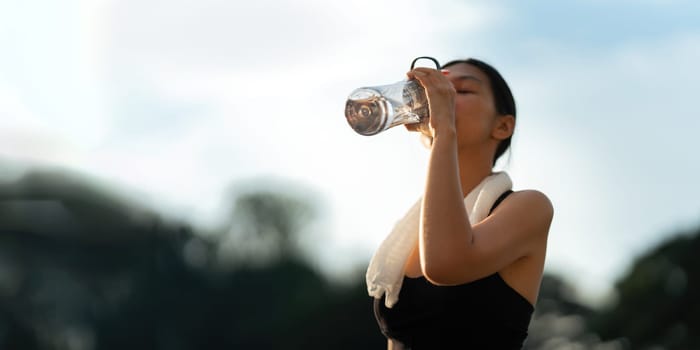 Beautiful young Asian woman stops to drinking water and wipe off her sweat after her evening run at park.