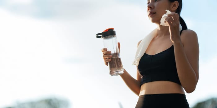 Beautiful young Asian woman stops to drinking water and wipe off her sweat after her evening run at park.