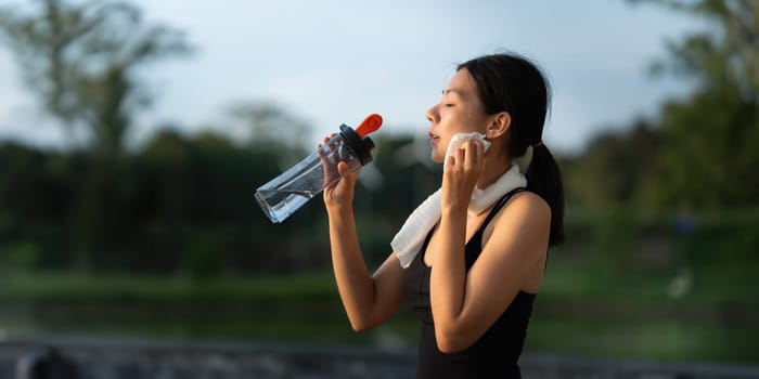 Beautiful young Asian woman stops to drinking water and wipe off her sweat after her evening run at park.
