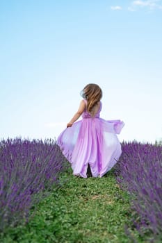 A young girl in a purple long dress runs between the lavender rows on the grass, the clear blue sky in the background.