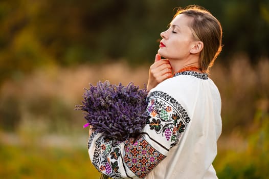 A young girl wearing a long embroidered shirt holds a lavender bouquet in her hands, photo at sunset.