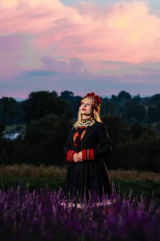 A girl in a chelsea headdress and a black dress decorated with red embroidery stands between lavender bushes.