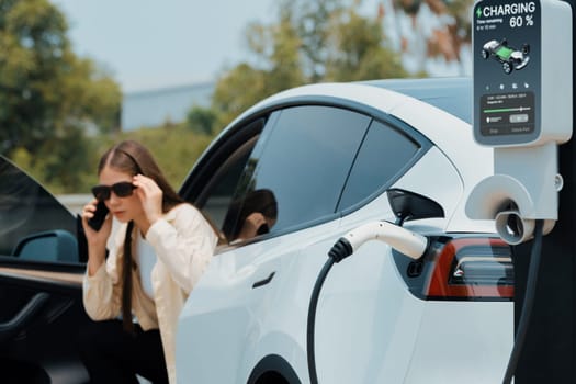 Young woman recharge her EV electric vehicle at green city park parking lot while talking on phone. Sustainable urban lifestyle for environment friendly EV car with battery charging station. Expedient