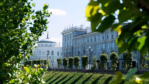 Central street of the city with adorable white houses and many green trees on blue cloudy sky background. Summer in the city, architecture concept.