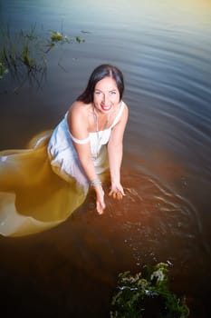 Adult mature brunette woman in a white dress, sundress and a wreath of flowers in summer in water of river or lake in evening at sunset. Celebration of the Slavic pagan holiday of Ivan Kupala