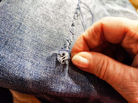 Woman repairing a pair of blue jeans. Fingers with a needle and torn jeans fabric. Sewing up a tear on blue fabric