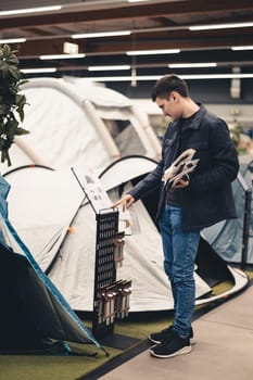 One young Caucasian man in a blue jacket stands sideways and reads the instructions for a tent in a store, close-up side view.