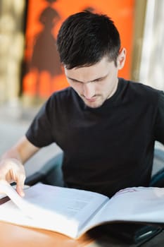 Portrait of one young handsome caucasian brunette man in a dark gray t-shirt sits at a table on the street terrace of a restaurant and looks through a book menu, close-up side view.