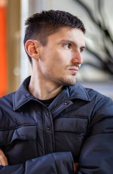 Portrait of one young handsome caucasian brunette guy in a dark fashionable jacket sitting at the table of the street terrace of the restaurant looking to the side, close-up side view.