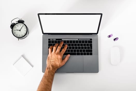 A man works at home at laptop on white table surrounded by an alarm clock, notebook and crystals for mental balance.