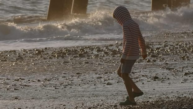 Boy walking by the sea, enjoying summer vibes. Creative. Boy child on a pebble beach in slow motion during sunset