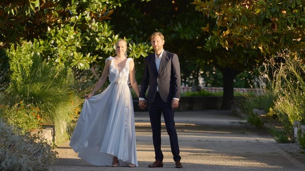 Bride and groom walking in the park. Action. A beautiful landscape with a green park where elegant newlyweds are walking by the hand. High quality 4k footage