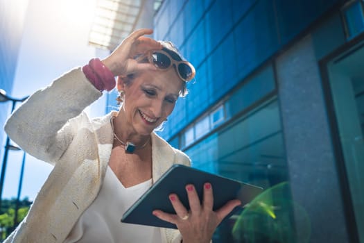 Mature business woman with technology happy working with a tablet holding sun glasses in a modern city. High quality photo