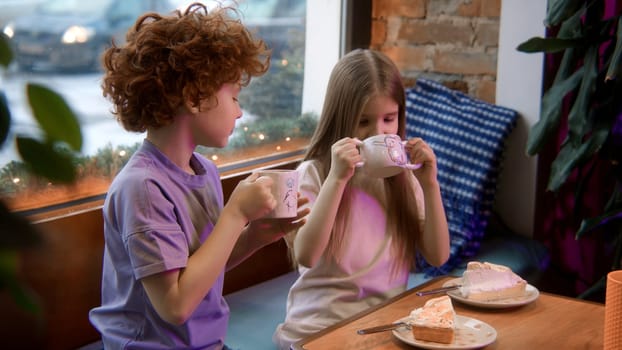 Two cute little kids having sweet meal in a cafe. Stock footage. Boy and girl holding cup of milk cocoa and looking happy