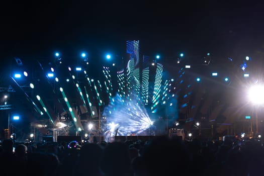 A music-filled night comes to life at a concert festival main event with a cheering unrecognizable crowd in front of the brightly lit stage. The lens flare adds to the fun and excitement.