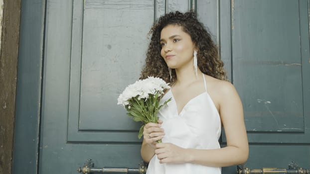 Beautiful young woman with bouquet of white flowers on street. Action. Attractive young woman smiles and holds bouquet of flowers. Smiling woman in dress and bouquet of flowers is waiting for date.