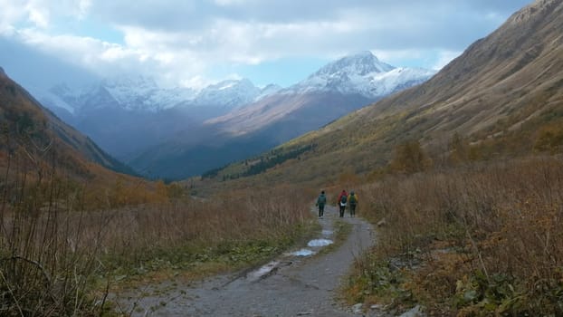 Tourists walk along trail in mountain valley in autumn. Creative. Hiking in mountain valley on cloudy autumn day. Trail in mountain valley with beautiful landscape of snowy peaks and cloudy sky.