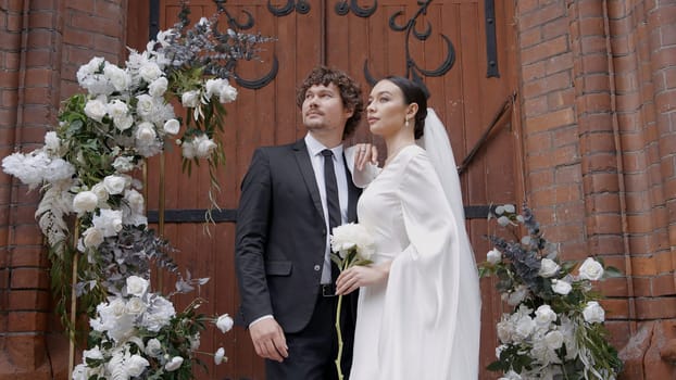 A bright and spectacular wedding couple. Action.Newlyweds where a bride in a white dress with a slit on her leg and a man with a beard in a suit walking next to a white arch with fresh flowers. High quality 4k footage