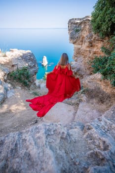 Red dress sea woman. Happy woman with flowing hair in a long flowing red dress stands on a rock near the sea. Travel concept, photo session at sea