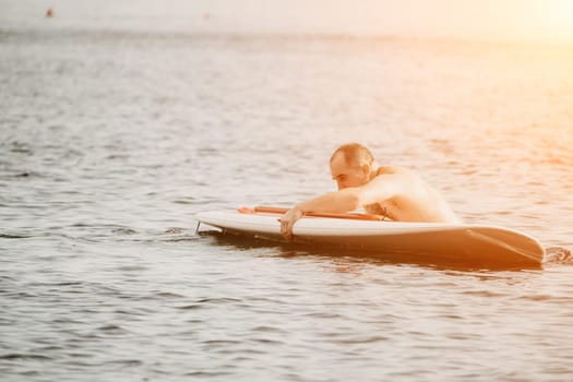 Active mature male paddler with his paddleboard and paddle on a sea at summer. Happy senior man stands with a SUP board. Stand up paddle boarding - outdor active recreation in nature