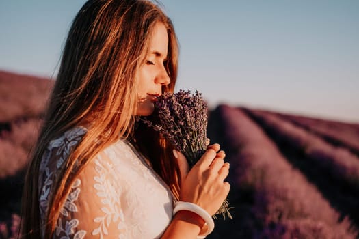 Close up portrait of young beautiful woman in a white dress and a hat is walking in the lavender field and smelling lavender bouquet.