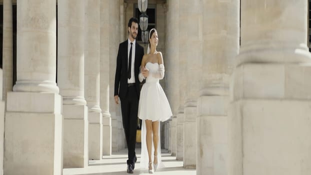 Lovely bride and groom walking along the columns in park. Action. Man in suit and a woman in short white dress near marble pillars