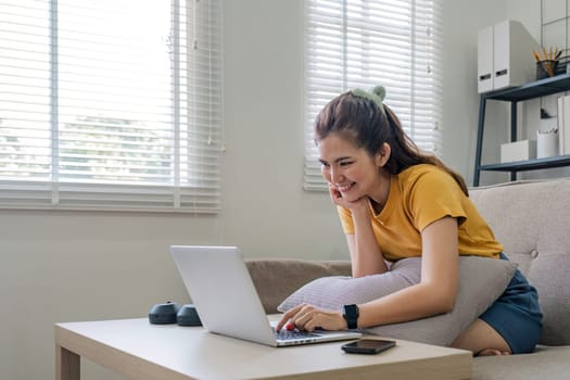 Beautiful Asian woman wearing casual clothes On the sofa using a laptop computer, entertaining with social media, relaxing, smiling and laughing..