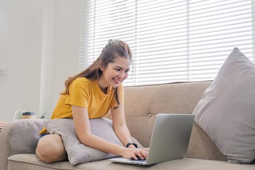 Beautiful Asian woman wearing casual clothes On the sofa using a laptop computer, entertaining with social media, relaxing, smiling and laughing..