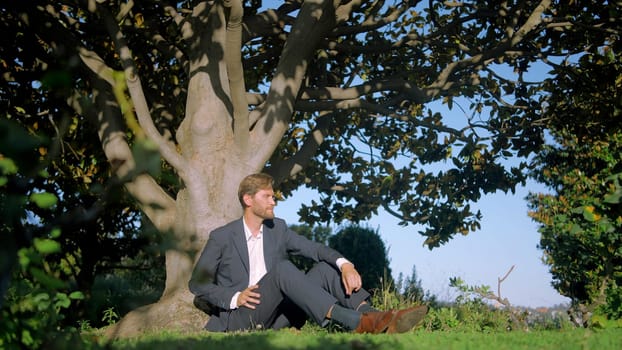 Man groom in suit sitting by the tree in the city park. Action. Young man having a rest under the tree