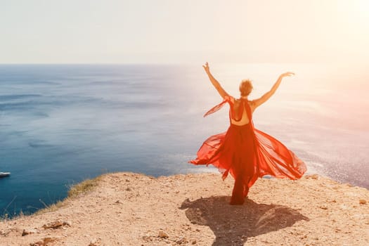 Side view a Young beautiful sensual woman in a red long dress posing on a rock high above the sea during sunrise. Girl on the nature on blue sky background. Fashion photo.