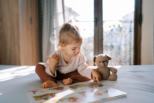 Little girl sits on a bed in a room and reads a book while guiding her finger across the page. High quality photo