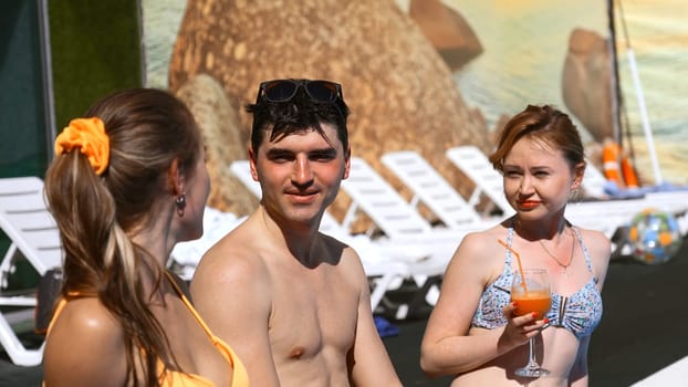 Three friends talking and having fun on the edge of the outdoors swimming pool. Clip. Two women and a man relaxing by the hotel pool