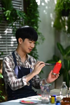 Happy Asian man painting on a glass jar with a brush in workshop. Indoors leisure activity concept.