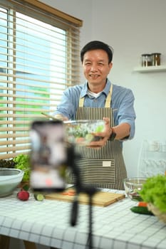 Cheerful senior male showing bowl of salad in front of smartphone while recording vlog video in kitchen