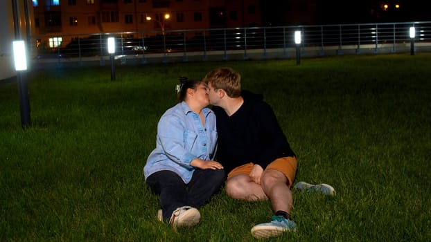 Happy loving couple sitting on green grass and making heart by folding hands. Media. Man and woman on green lawn near modern house