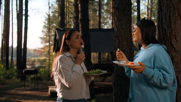 Barbecue on a summer day in coniferous forest. Stock footage. Two adult girls standing and communicating while eating