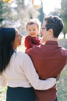 Smiling mom hugs dad with a little girl in his arms. Back view. High quality photo