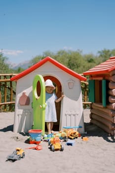 Little girl comes out of a toy house and looks at the toys on the sand. High quality photo