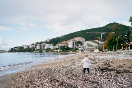 Little girl walks along the pebbly seashore against the backdrop of mountains and apartment buildings. Back view. High quality photo